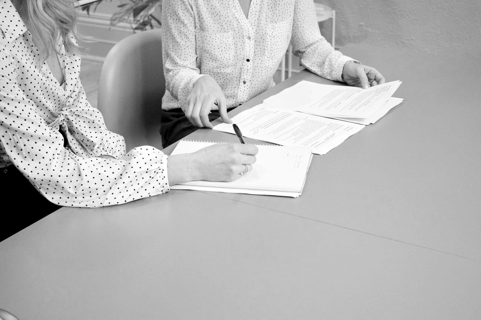 Duas pessoas à mesa: uma anota num caderno enquanto a outra aponta um trecho do contrato