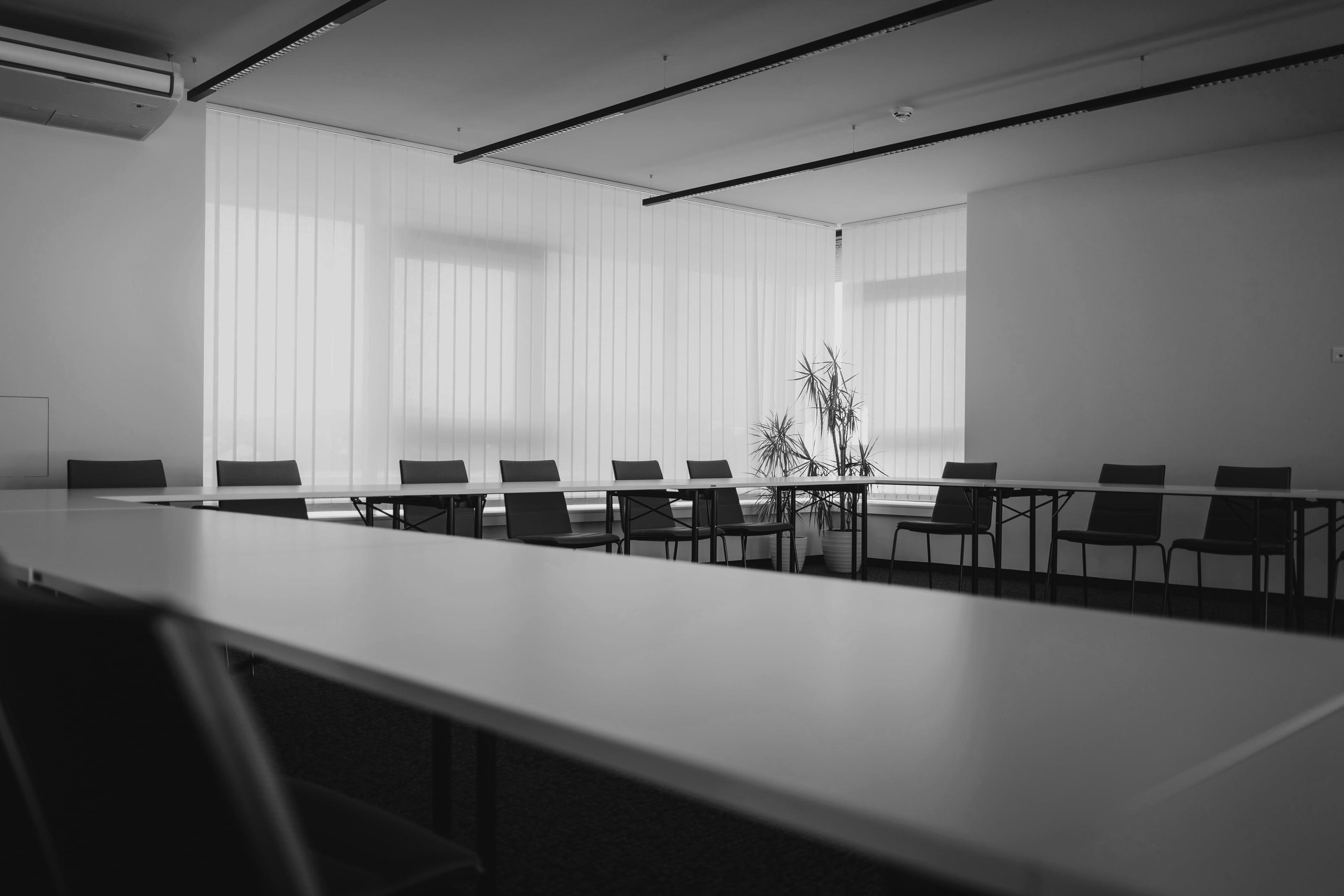 Sala de reuniões vazia, com a mesa em U e as persianas fechadas filtrando a luz da manhã