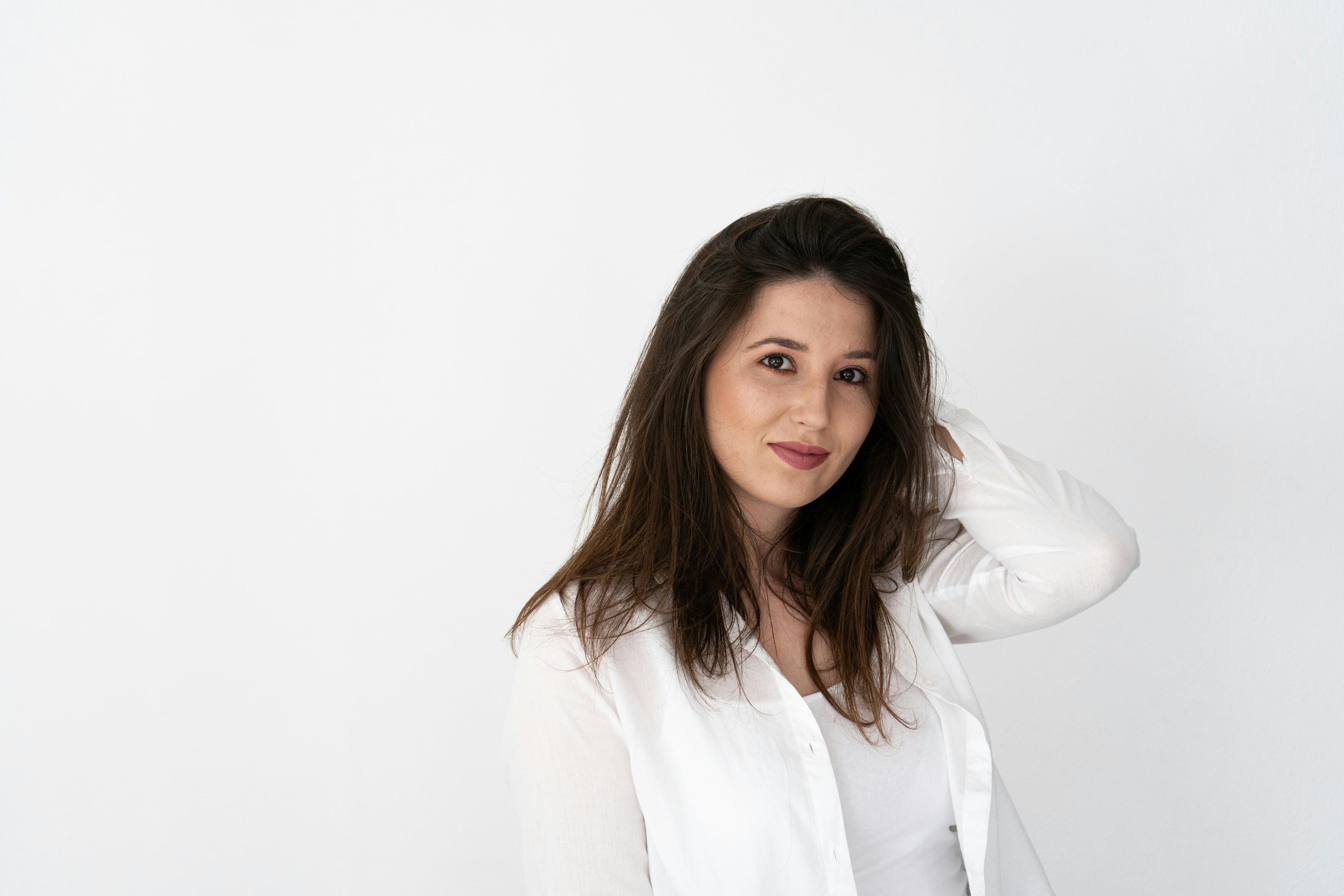 Mulher de cabelo castanho e camisa branca diante de uma parede clara, com a mão no cabelo