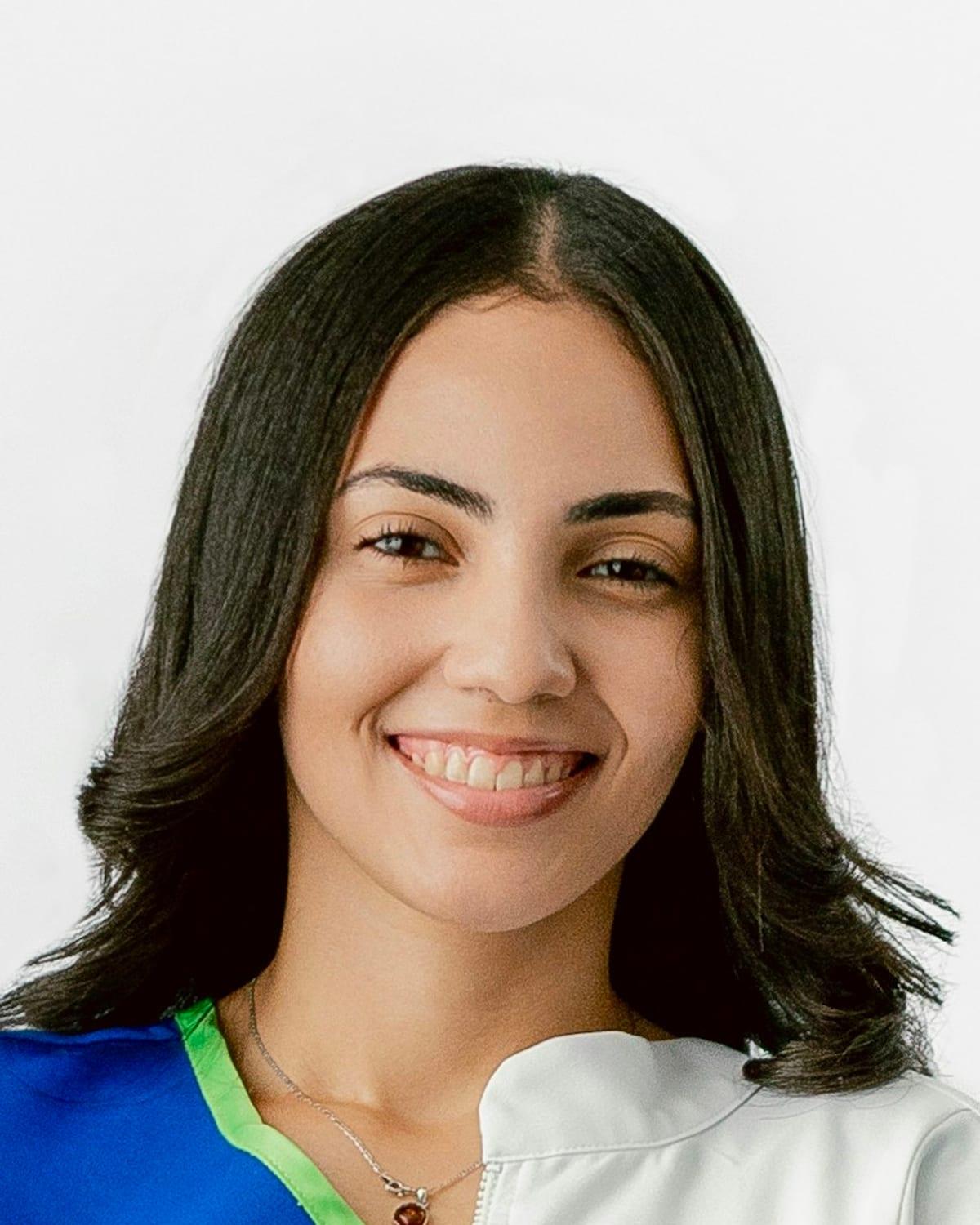 Retrato de Helena Prado sorrindo, de jaleco branco sobre o pijama cirúrgico azul, diante de uma parede clara