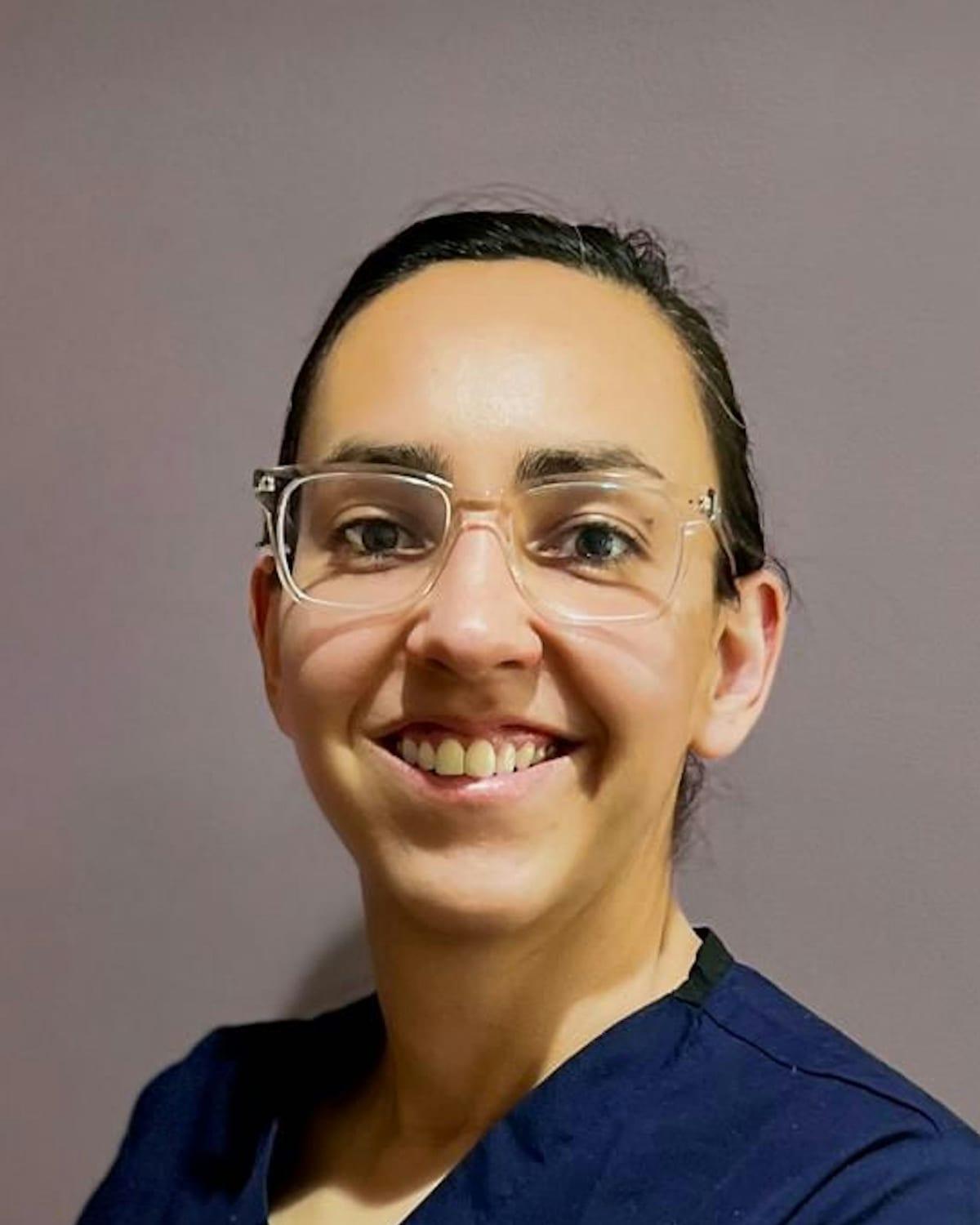 Retrato de Lúcia Tavares de óculos e uniforme azul-marinho, sorrindo, diante de uma parede lilás acinzentada