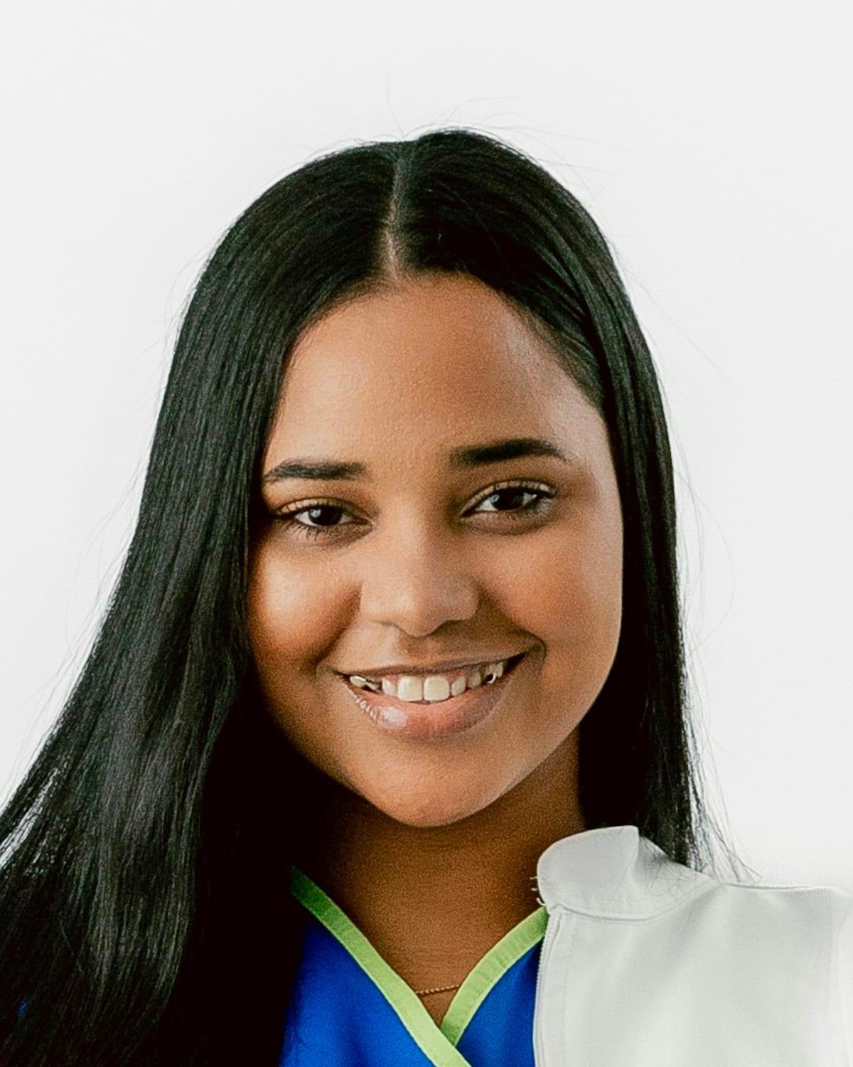 Retrato de Rafaela Nunes sorrindo, de jaleco branco sobre o pijama cirúrgico azul, diante de uma parede clara