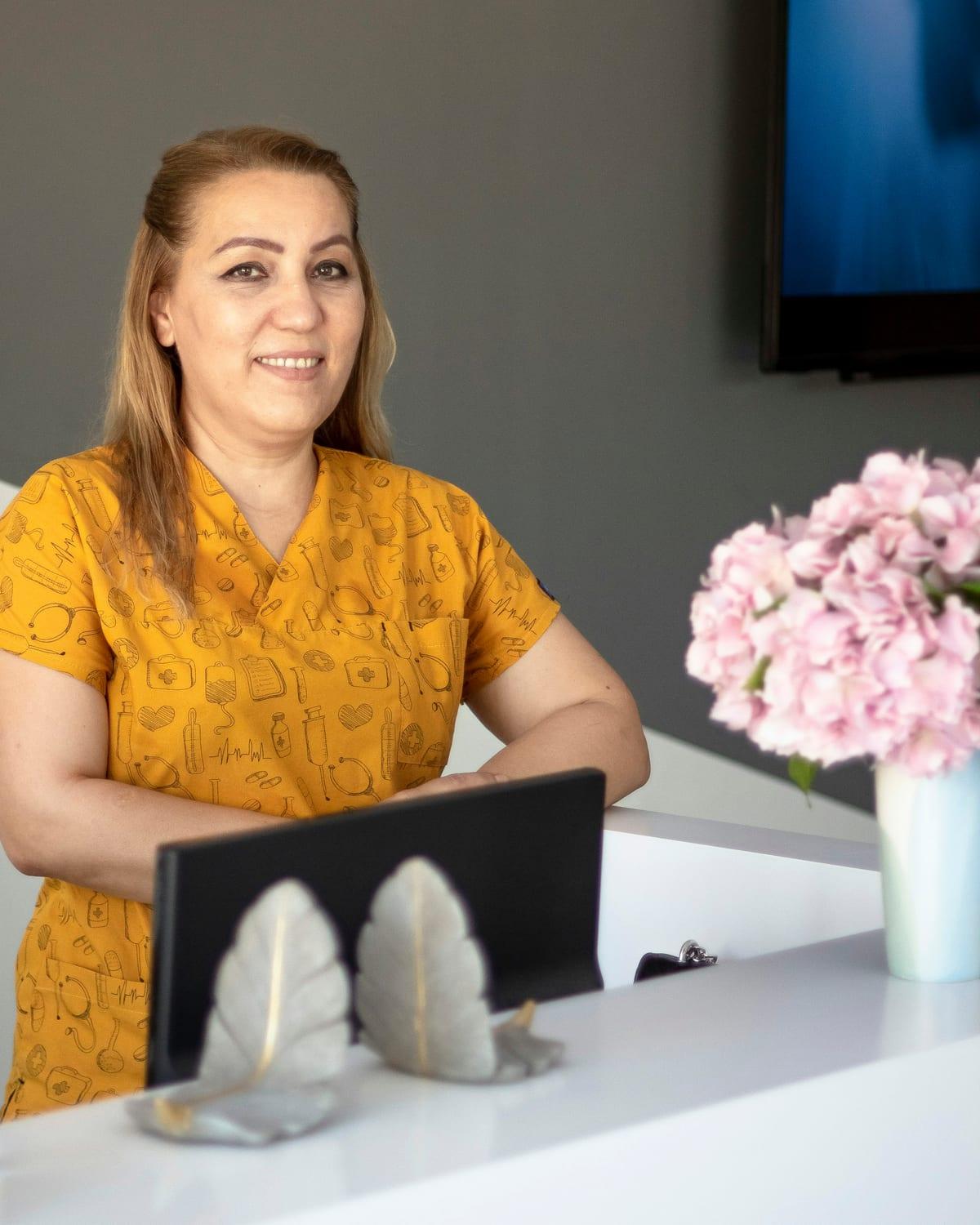 Recepcionista de uniforme estampado sorrindo atrás do balcão branco, ao lado de um vaso de hortênsias