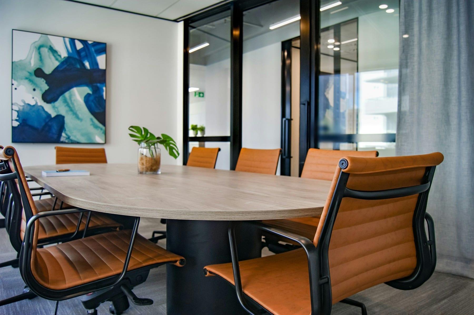 Sala de reuniões com mesa de madeira clara, cadeiras de couro caramelo e divisórias de vidro