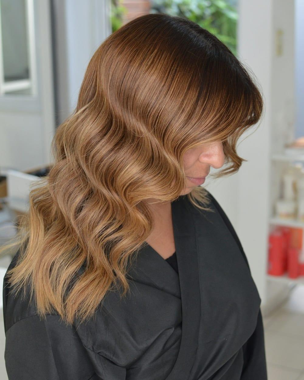 Cliente de capa preta no salão, com cabelo castanho escuro na raiz e ondas cor de caramelo nas pontas.