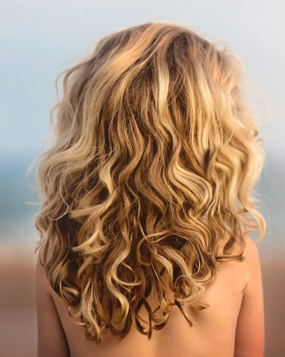 Cabelo cacheado e volumoso visto de costas, com mechas loiro dourado misturadas ao castanho.