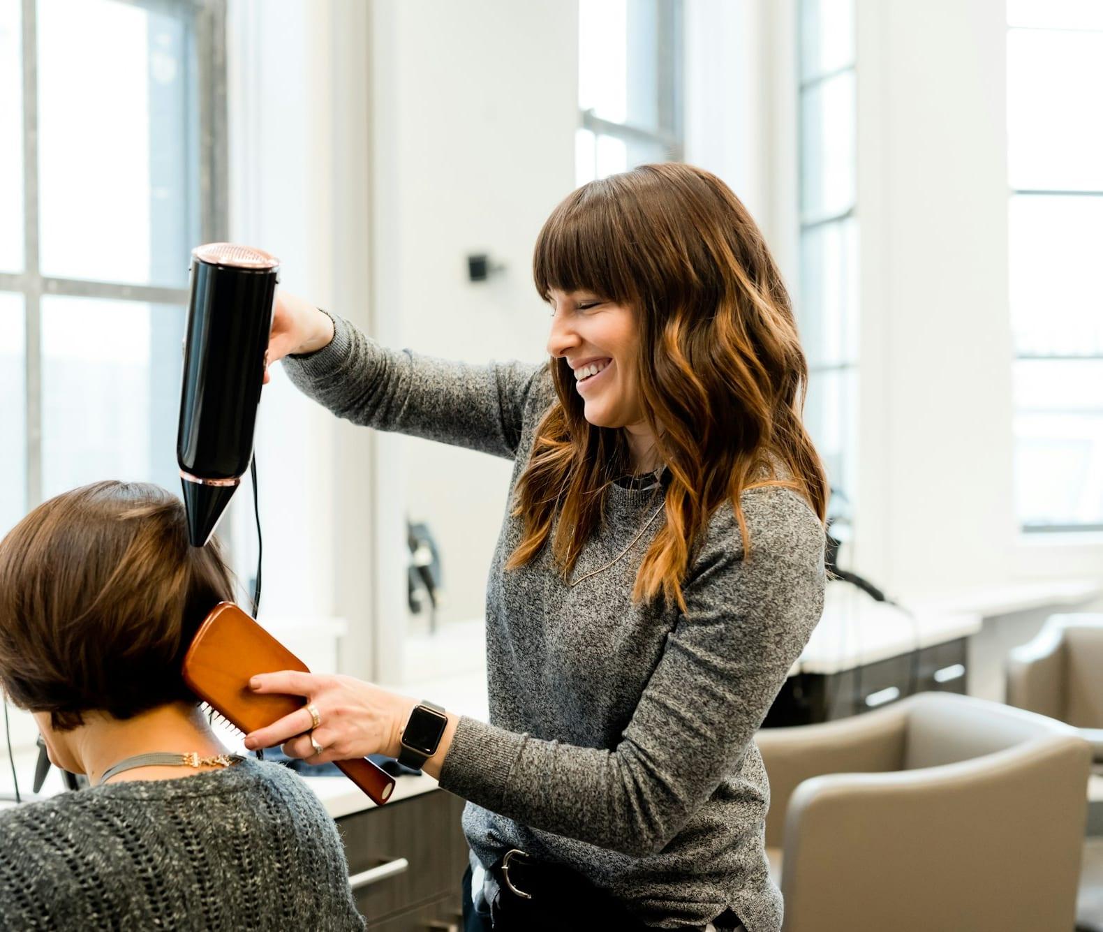 Tati Roveda, de franja e suéter cinza, sorri enquanto seca com escova o cabelo curto de uma cliente no salão.
