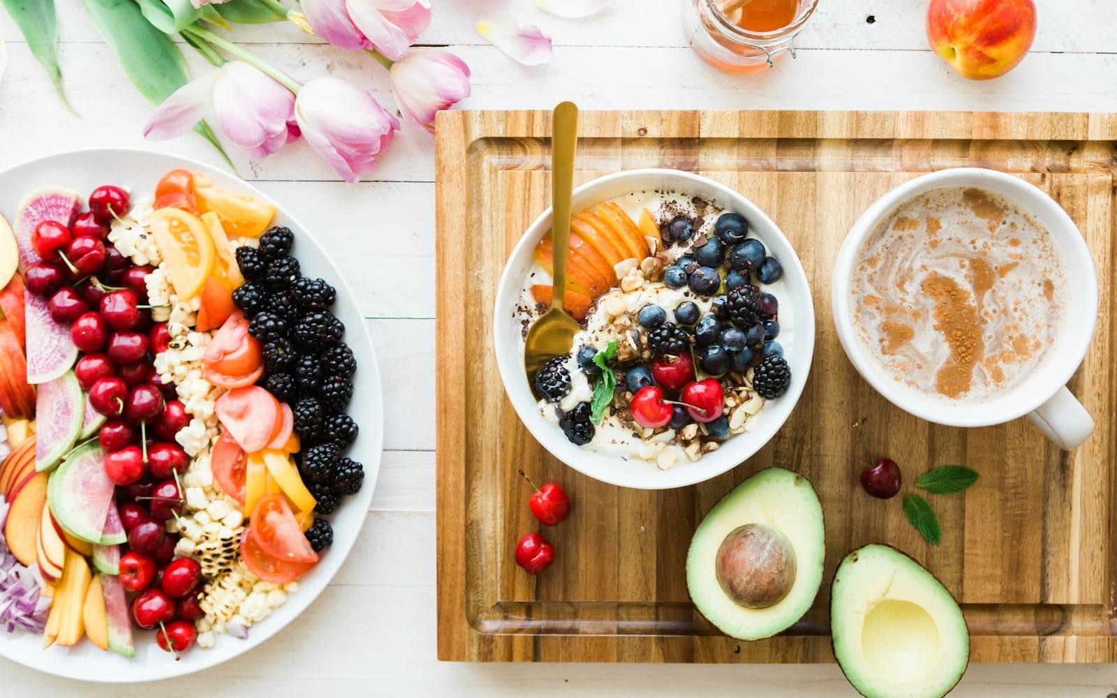 Tigela de iogurte com aveia, mirtilo, amora e cereja numa tábua, ao lado de café com leite, abacate aberto e um prato de frutas