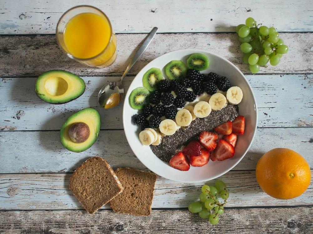 Tigela de frutas com kiwi, banana, amora e morango, pão integral, abacate e suco de laranja sobre mesa de madeira