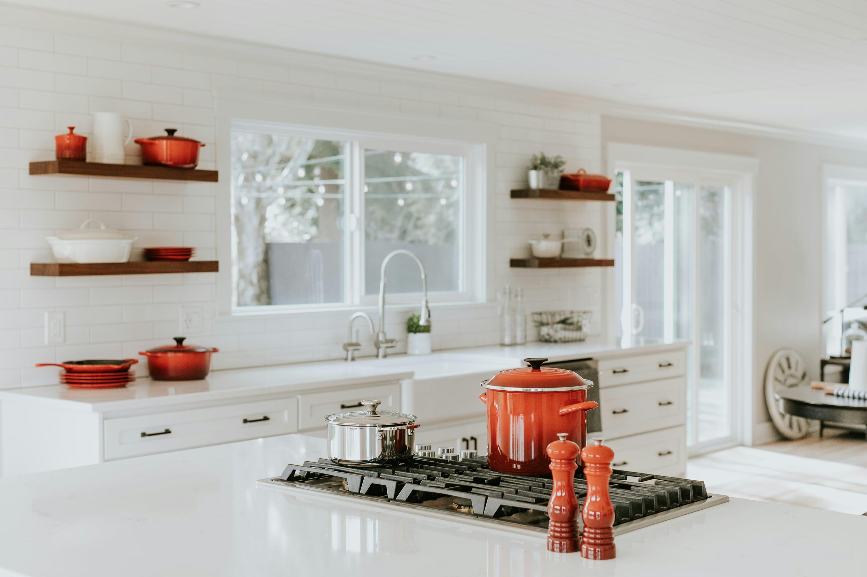 Cozinha branca com caldeirão vermelho e caçarola de inox no cooktop, moedores de pimenta e prateleiras com panelas vermelhas