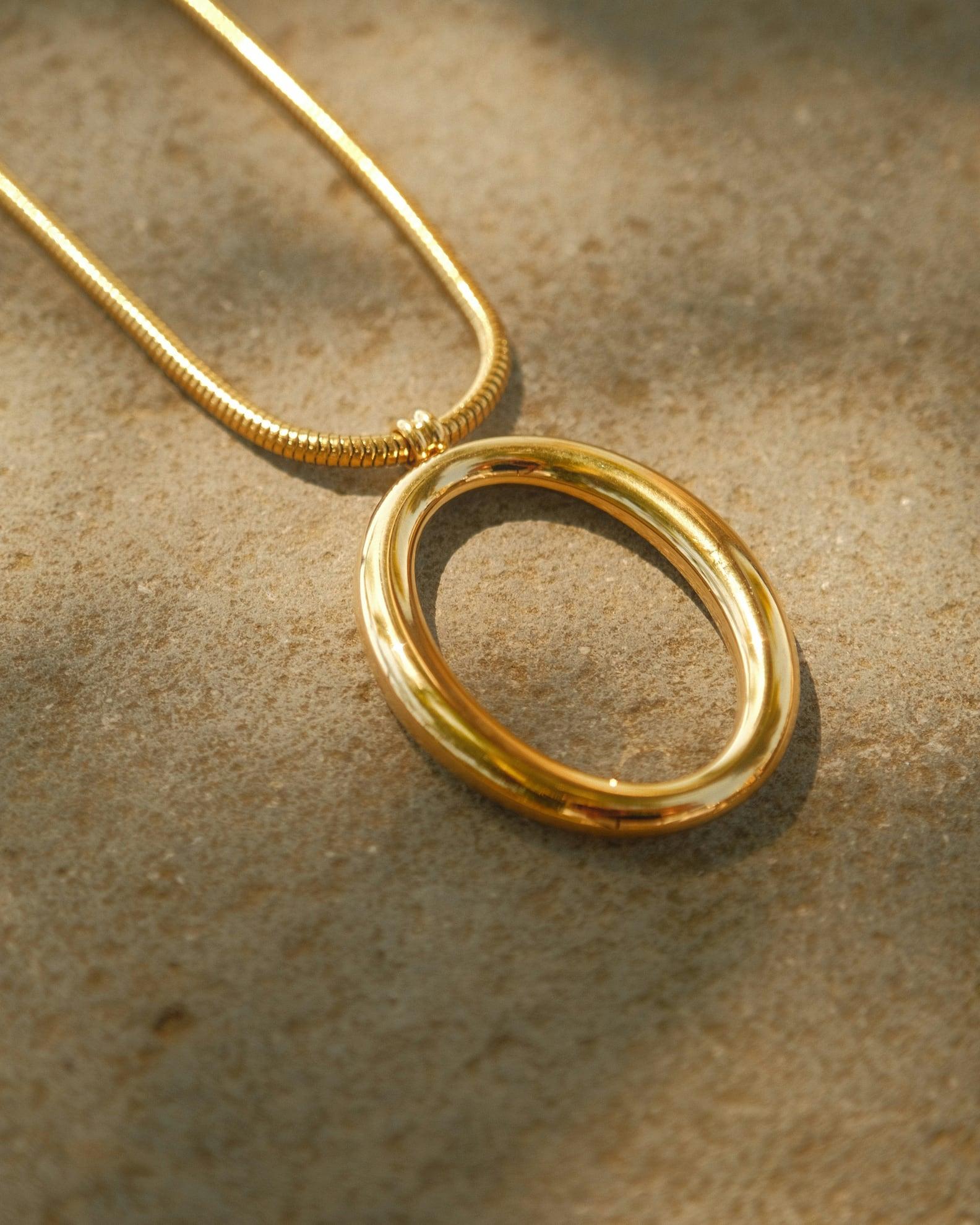Colar dourado com pingente oval vazado, apoiado sobre areia