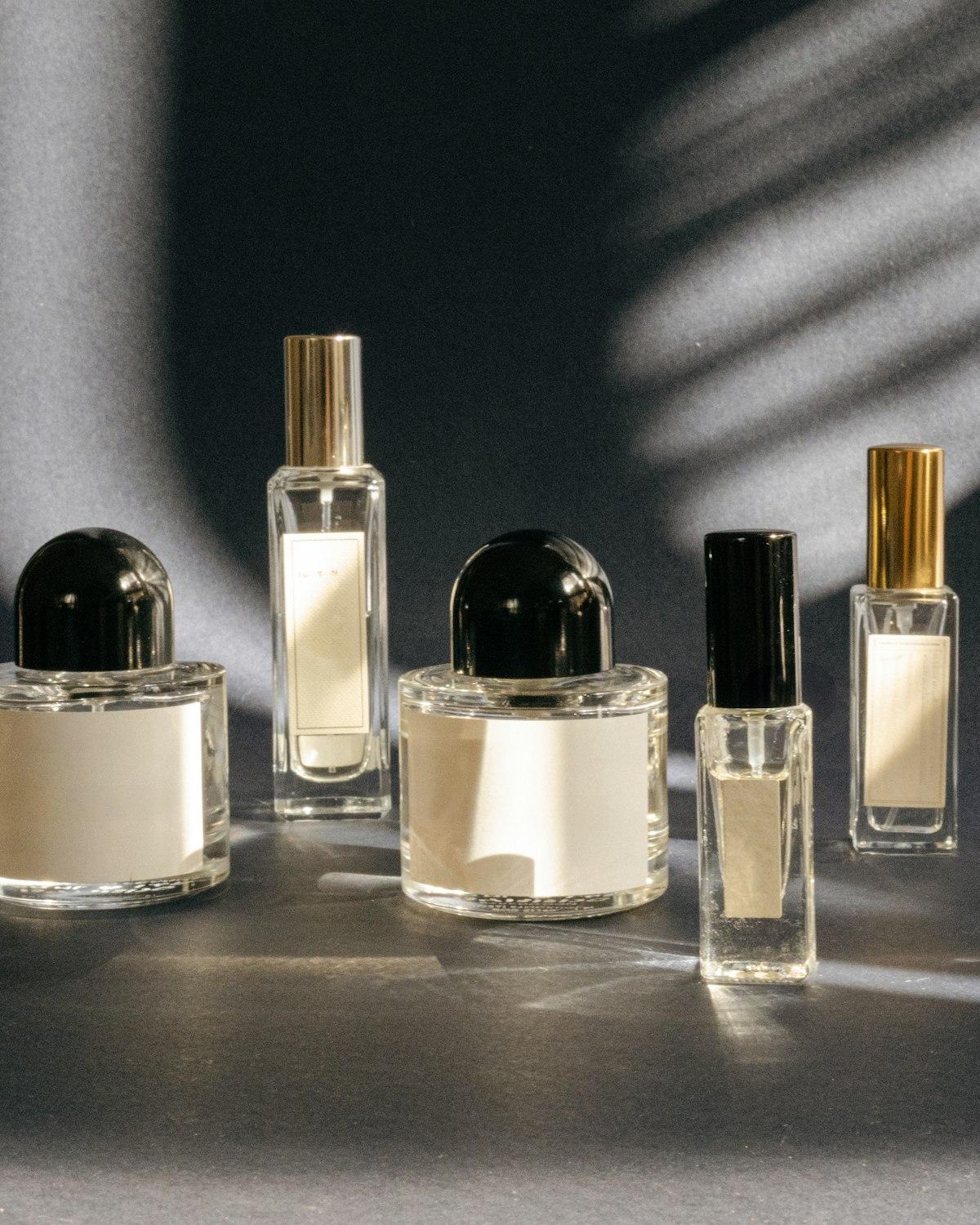 Cinco frascos de perfume com rótulos em branco, de tampas pretas e douradas, sob luz recortada sobre fundo escuro